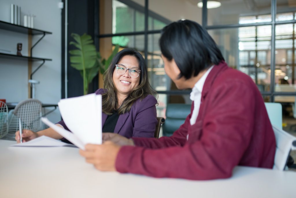Smiling Asian woman sitting at a desk with a man holding documents