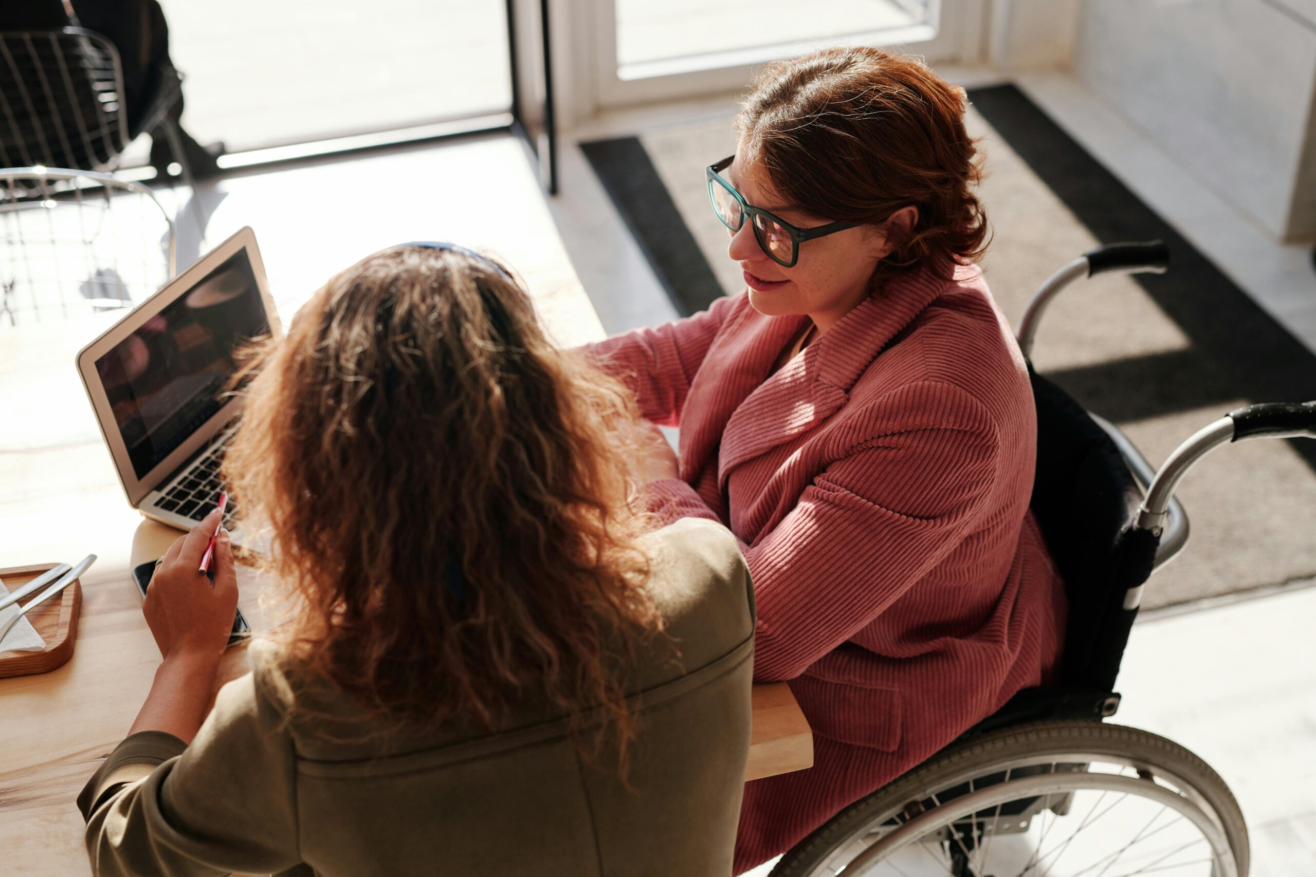 women-in-wheelchair-sitting-at-desk-talking-to-colleague A manager and an employee having a one-on-one meeting